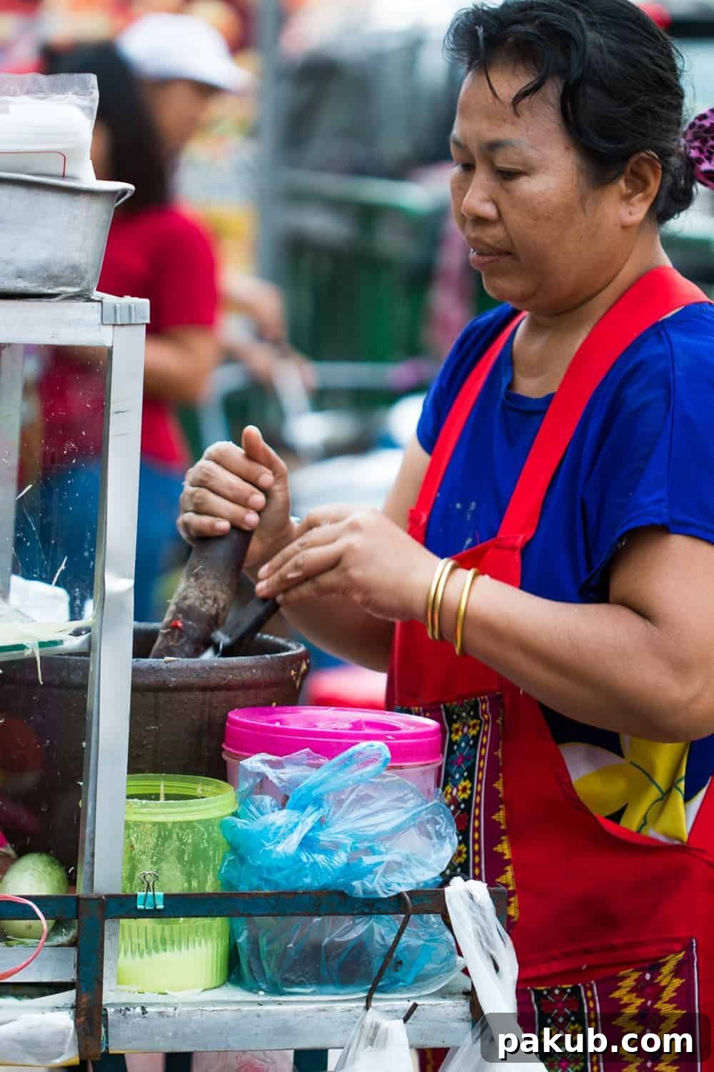 A dedicated woman, dressed in a practical blue outfit and a red apron, intently cooking a flavorful street food dish over a hot wok in a bustling Bangkok market
