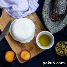 A beautifully arranged photograph of homemade cream cheese on a wooden board, accompanied by olive oil, fresh herbs, and a plum, showcasing serving suggestions.