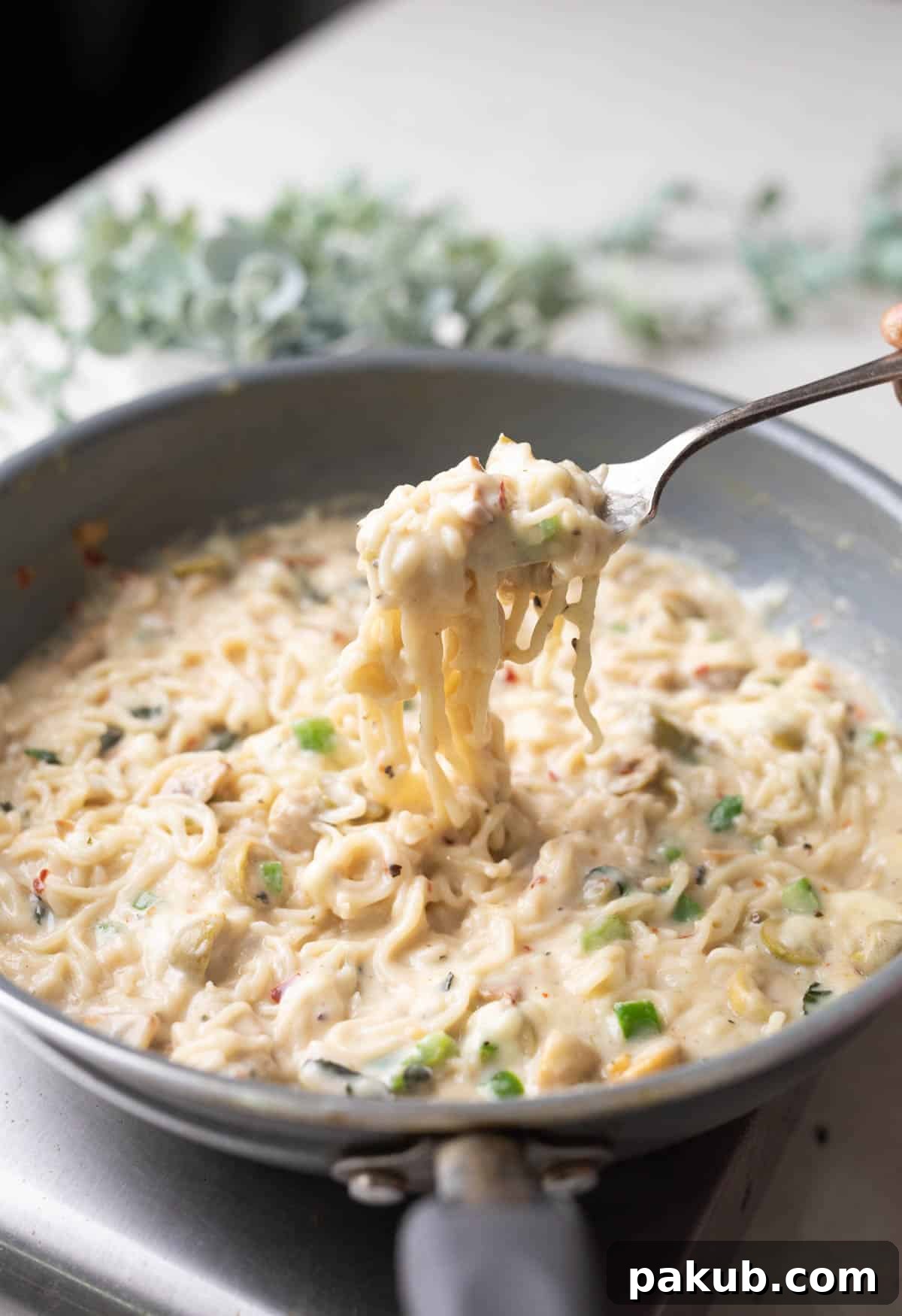 A close-up shot of creamy Italian Alfredo Maggi noodles in a grey pan, with a fork lifting a portion, showcasing the rich sauce and mushrooms.