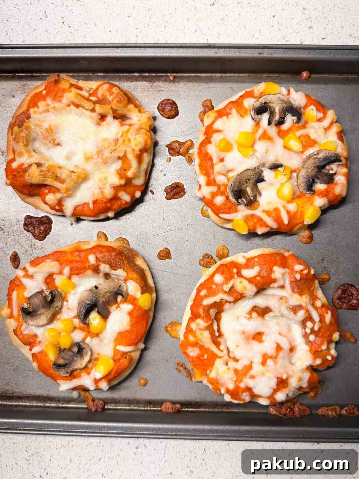 Kid-Friendly Mini Veggie Pizzas 3 Four delicious homemade mini vegetarian pizzas on a dark baking sheet, adorned with mushrooms, corn, and melted cheese, ready to be enjoyed.